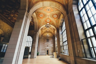 An elegant interior with high arched ceilings adorned with intricate patterns and chandeliers. Large windows allow ample natural light to illuminate the spacious area with polished wooden flooring. Architectural details create a classical and grand atmosphere.