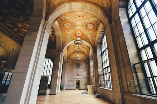 An elegant interior with high arched ceilings adorned with intricate patterns and chandeliers. Large windows allow ample natural light to illuminate the spacious area with polished wooden flooring. Architectural details create a classical and grand atmosphere.