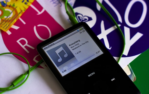 A black portable music player with a screen displaying a song titled 'Kreuzberg' by Bloc Party from the album 'Weekend In The City'. Green earphones are connected to the device. Surrounding the player are colorful pieces of paper with bold text and designs.