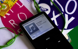 A black portable music player with a screen displaying a song titled 'Kreuzberg' by Bloc Party from the album 'Weekend In The City'. Green earphones are connected to the device. Surrounding the player are colorful pieces of paper with bold text and designs.