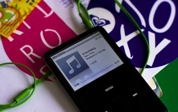 A black portable music player with a screen displaying a song titled 'Kreuzberg' by Bloc Party from the album 'Weekend In The City'. Green earphones are connected to the device. Surrounding the player are colorful pieces of paper with bold text and designs.