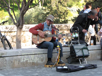 A musician playing an acoustic guitar connected to a compact MusicCube amp on a sunny park bench.