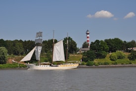 A sailing ship with tall masts and several sails is depicted on calm waters near a green, wooded shoreline. A distinct lighthouse with red and white stripes stands prominently in the background, surrounded by greenery and a few buildings.