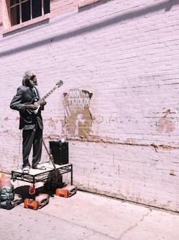 An older man with a gray beard stands on a makeshift platform, playing an electric guitar. He is dressed in a black suit with sandals and is positioned against a weathered brick wall with a partially torn poster. Several orange power tools are on the ground beneath him.