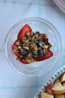 A glass dish filled with layers of yogurt, granola, strawberries, and blueberries sits on a white and blue patterned surface. The yogurt provides a creamy base, topped with vibrant red strawberries and dark blue blueberries. A layer of crunchy granola is sprinkled on top, adding texture. Adjacent to the dish, there is a plate with slices of apples, captured partially in the frame.