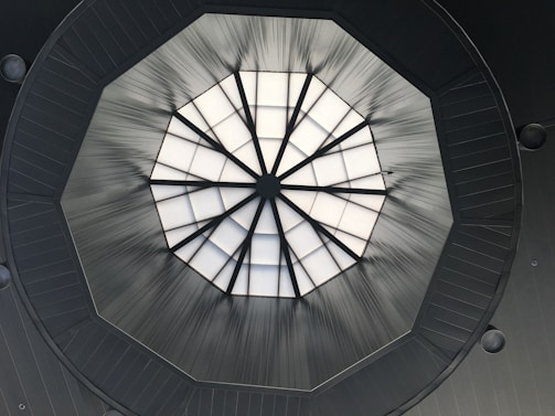 A geometric skylight with a domed structure, segmented into triangular glass panes. The light is diffused, creating a pattern of radiating lines against the dark framework surrounding the glass. The clean lines and angular design give a modern architectural feel.