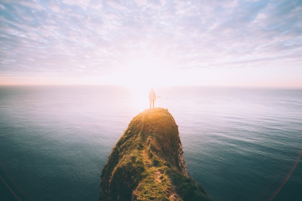 A golden hour shot of a traveler standing on a cliff overlooking a vast ocean under a navy-blue sky.