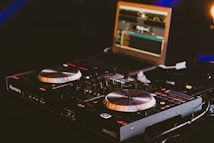 A DJ controller featuring numerous knobs, buttons, and dual jog wheels is prominently displayed. In the background, a laptop with music software running on its screen is visible, adding to the setup's functionality. The lighting is dim, creating an ambient atmosphere with a focus on the equipment.