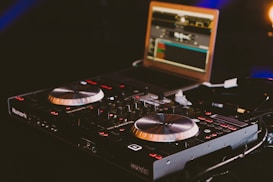 A DJ controller featuring numerous knobs, buttons, and dual jog wheels is prominently displayed. In the background, a laptop with music software running on its screen is visible, adding to the setup's functionality. The lighting is dim, creating an ambient atmosphere with a focus on the equipment.