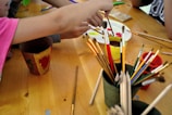 A group of children or people engaged in a painting activity, with various paintbrushes and containers on a wooden table. Bright colors are visible on a palette and there are hand movements indicating active painting.