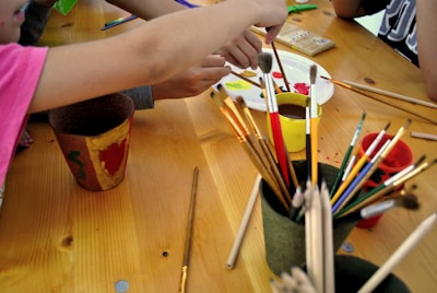 A group of children or people engaged in a painting activity, with various paintbrushes and containers on a wooden table. Bright colors are visible on a palette and there are hand movements indicating active painting.