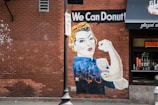A mural on a red brick wall depicting a strong woman in the style of the iconic Rosie the Riveter poster, holding a donut. The text 'We Can Donut!' appears above her. She wears a blue shirt and a yellow polka-dot bandana. There's a storefront with neon lights and donut decorations nearby. A streetlamp and 'Right Turn Only' sign are also visible.