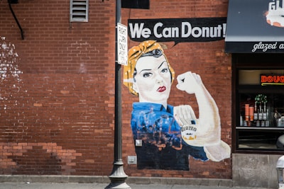 A mural on a red brick wall depicting a strong woman in the style of the iconic Rosie the Riveter poster, holding a donut. The text 'We Can Donut!' appears above her. She wears a blue shirt and a yellow polka-dot bandana. There's a storefront with neon lights and donut decorations nearby. A streetlamp and 'Right Turn Only' sign are also visible.