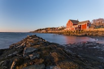 A coastal landscape featuring a red barn-style house on stilts situated by a rocky shoreline. The house overlooks a calm body of water under a clear blue sky transitioning to sunset. The shoreline is covered in algae and rocks, leading the eye to the distance where a small community of houses is nestled among a line of trees.