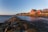 A coastal landscape featuring a red barn-style house on stilts situated by a rocky shoreline. The house overlooks a calm body of water under a clear blue sky transitioning to sunset. The shoreline is covered in algae and rocks, leading the eye to the distance where a small community of houses is nestled among a line of trees.