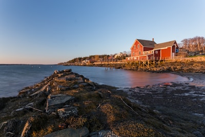A coastal landscape featuring a red barn-style house on stilts situated by a rocky shoreline. The house overlooks a calm body of water under a clear blue sky transitioning to sunset. The shoreline is covered in algae and rocks, leading the eye to the distance where a small community of houses is nestled among a line of trees.