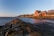A coastal landscape featuring a red barn-style house on stilts situated by a rocky shoreline. The house overlooks a calm body of water under a clear blue sky transitioning to sunset. The shoreline is covered in algae and rocks, leading the eye to the distance where a small community of houses is nestled among a line of trees.