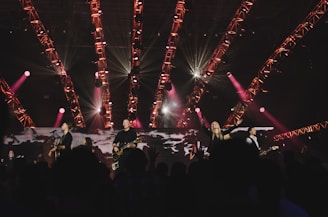 A band is performing live on stage with various musical instruments under a structure of brightly lit, red and pink stage lights. The crowd is visible in the foreground, enjoying the performance, with some people raising their hands. The atmosphere is vibrant and energetic.