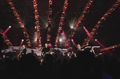 A band is performing live on stage with various musical instruments under a structure of brightly lit, red and pink stage lights. The crowd is visible in the foreground, enjoying the performance, with some people raising their hands. The atmosphere is vibrant and energetic.
