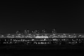 A black and white image capturing a night scene of a railway station with bright artificial lights. In the background, several high-rise buildings loom against the dark sky. The station name and some signage are illuminated with white lights, providing contrast against the night.