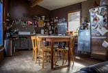 A rustic kitchen with wooden furniture and various kitchen utensils scattered around. The walls are adorned with shelves holding jars and cookware, and the table in the center is surrounded by chairs. Sunlight streams in through a window, illuminating a refrigerator covered with photos and notes.