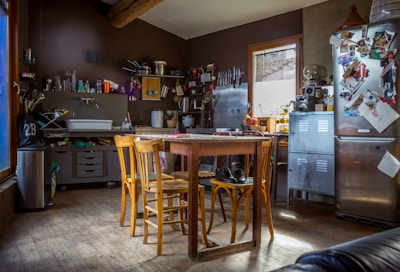 A rustic kitchen with wooden furniture and various kitchen utensils scattered around. The walls are adorned with shelves holding jars and cookware, and the table in the center is surrounded by chairs. Sunlight streams in through a window, illuminating a refrigerator covered with photos and notes.
