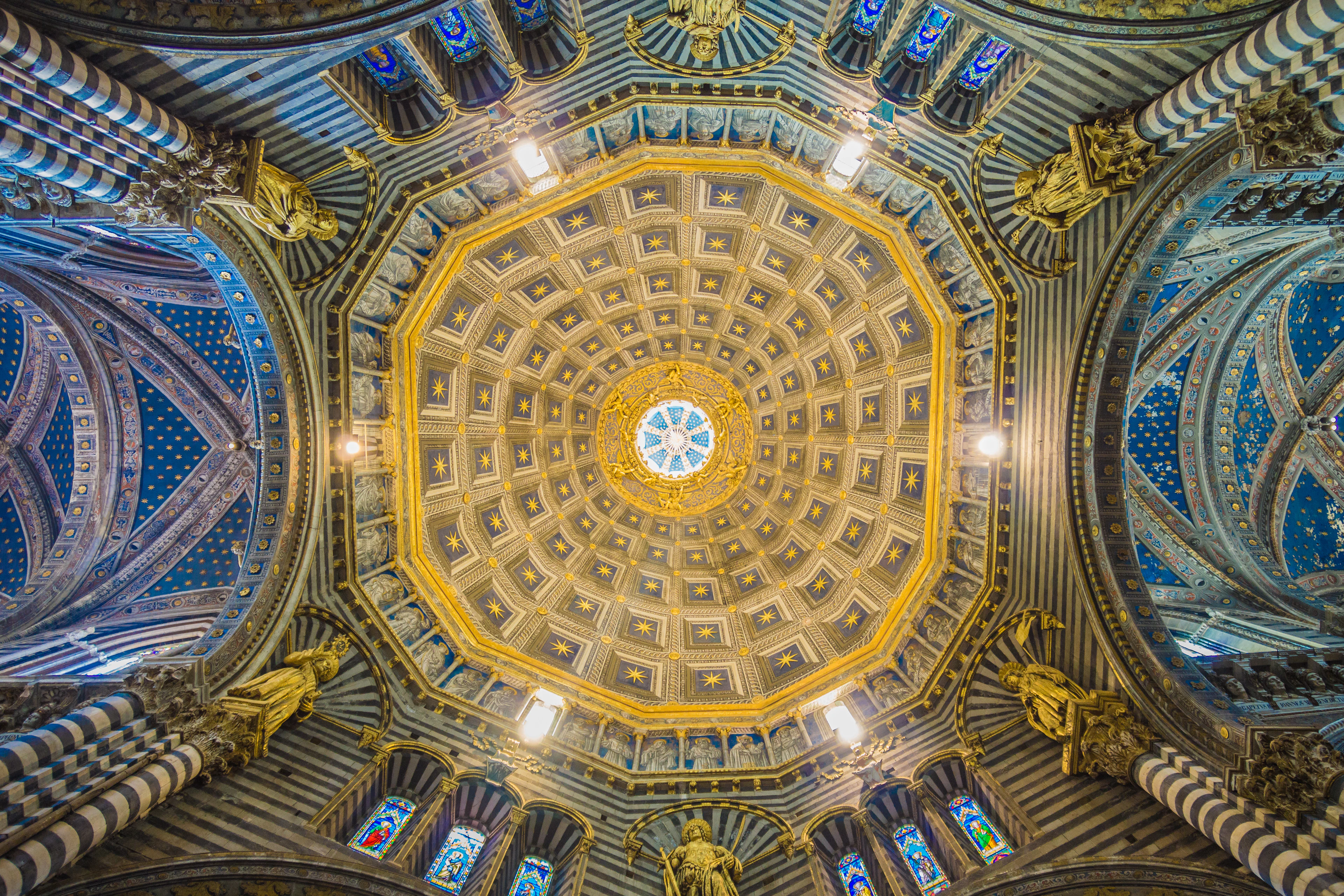 Looking straight up into the gilded coffered dome of Siena Cathedral, its gold hexagon panels and painted stars ringed by deep blue vaults, stained glass and black-and-white striped marble piers.