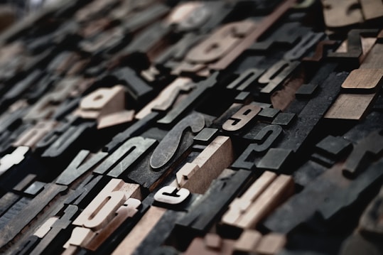 A close-up view of vintage wooden letterpress type blocks arranged randomly. The image captures an assortment of letters and punctuation marks of various sizes and fonts, creating an intricate and textured pattern.