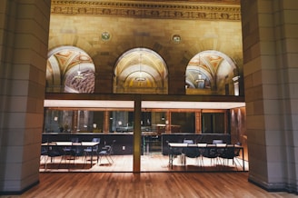 A spacious and elegant interior featuring arched ceilings with ornate designs, grand chandeliers, and expansive windows. The space includes a row of tables and chairs, suggesting a formal or conference setting. The flooring is polished wood, complemented by the warm lighting.