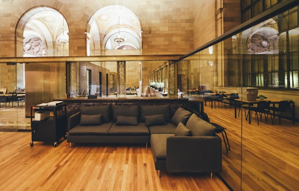 A spacious interior with a mix of classical and modern design elements. The room features arched ceilings with ornate details, large reflective glass windows on one side, and a polished wooden floor. A sleek dark sofa with matching pillows is positioned in the foreground, accompanied by a mobile cart with office equipment. In the background, tables and chairs are arranged neatly in a semi-formal setting.