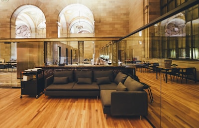A spacious interior with a mix of classical and modern design elements. The room features arched ceilings with ornate details, large reflective glass windows on one side, and a polished wooden floor. A sleek dark sofa with matching pillows is positioned in the foreground, accompanied by a mobile cart with office equipment. In the background, tables and chairs are arranged neatly in a semi-formal setting.