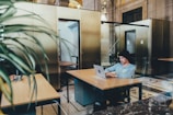 A person wearing a beanie is working on a laptop at a wooden table in a modern, open-space office. The area features high ceilings and is surrounded by glass partitions and minimalistic decor. A plant partially obscures the foreground, adding a touch of green.