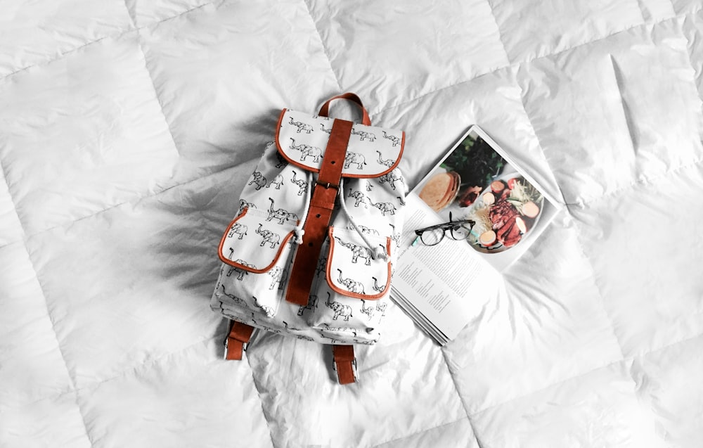 A white backpack on a bed mattress next to a cookbook and a pair of glasses