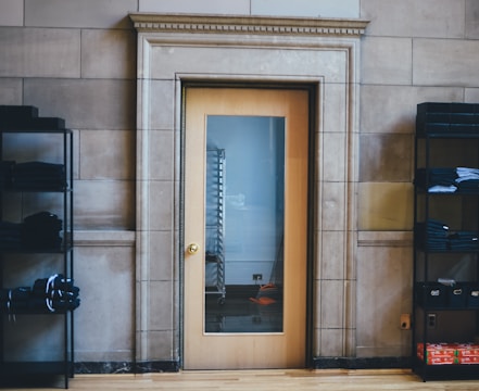 A wooden door with a glass panel in a stone wall, flanked by black metal shelving units holding neatly folded clothes. The door reflects a metal rack through the glass.