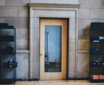 A wooden door with a glass panel in a stone wall, flanked by black metal shelving units holding neatly folded clothes. The door reflects a metal rack through the glass.