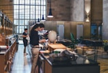 Inside a spacious cafe or restaurant with high ceilings and large windows, two people are engaged in various activities near a counter, which is stocked with items. The decor includes modern lighting fixtures, plants, and tables arranged in the background for seating.