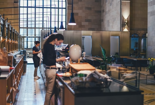Inside a spacious cafe or restaurant with high ceilings and large windows, two people are engaged in various activities near a counter, which is stocked with items. The decor includes modern lighting fixtures, plants, and tables arranged in the background for seating.