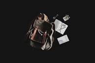 A rustic leather backpack is placed against a dark background, accompanied by a variety of travel-related items. Next to the backpack, there are a journal with handwritten notes, a pen, a passport, loose coins, a small book titled 'El Despertario,' and a neatly rolled-up cord or cable.