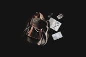 A rustic leather backpack is placed against a dark background, accompanied by a variety of travel-related items. Next to the backpack, there are a journal with handwritten notes, a pen, a passport, loose coins, a small book titled 'El Despertario,' and a neatly rolled-up cord or cable.