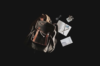A rustic leather backpack is placed against a dark background, accompanied by a variety of travel-related items. Next to the backpack, there are a journal with handwritten notes, a pen, a passport, loose coins, a small book titled 'El Despertario,' and a neatly rolled-up cord or cable.