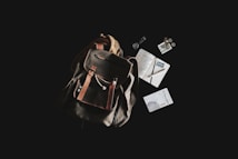 A rustic leather backpack is placed against a dark background, accompanied by a variety of travel-related items. Next to the backpack, there are a journal with handwritten notes, a pen, a passport, loose coins, a small book titled 'El Despertario,' and a neatly rolled-up cord or cable.