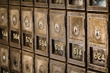 Rows of vintage-style post office boxes featuring ornate designs and numbered locks built into a wall. The metal boxes have a brass or bronze appearance with intricate patterns surrounding the locks.
