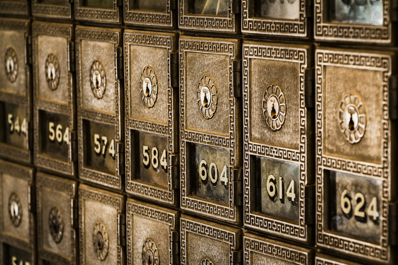 A close-up angled wall of ornate brass/bronze bank safe-deposit boxes with decorative keyholes and engraved numbers (544, 564, 574, 584, 604, 614