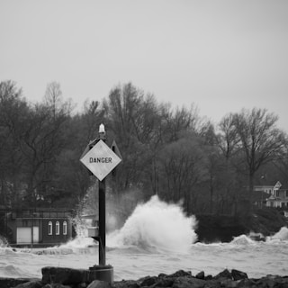 A monochrome image featuring a 'Danger' sign prominently in the foreground, set against a backdrop of a turbulent body of water with waves crashing ashore. The background includes bare trees and a structure with windows, likely a building or house, contributing to the sense of a stormy environment.