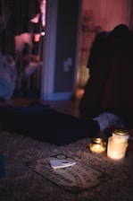 The image features a dimly lit room with a mysterious ambiance. A Ouija board is on a plush carpet, surrounded by three lit candles that provide a soft, warm glow. In the background, hanging beads create a partition between spaces, while various objects and clothing create an informal and slightly cluttered atmosphere.