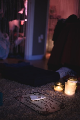 The image features a dimly lit room with a mysterious ambiance. A Ouija board is on a plush carpet, surrounded by three lit candles that provide a soft, warm glow. In the background, hanging beads create a partition between spaces, while various objects and clothing create an informal and slightly cluttered atmosphere.