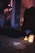 The image features a dimly lit room with a mysterious ambiance. A Ouija board is on a plush carpet, surrounded by three lit candles that provide a soft, warm glow. In the background, hanging beads create a partition between spaces, while various objects and clothing create an informal and slightly cluttered atmosphere.
