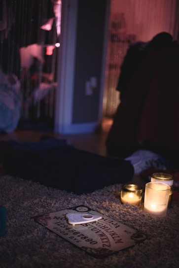 The image features a dimly lit room with a mysterious ambiance. A Ouija board is on a plush carpet, surrounded by three lit candles that provide a soft, warm glow. In the background, hanging beads create a partition between spaces, while various objects and clothing create an informal and slightly cluttered atmosphere.