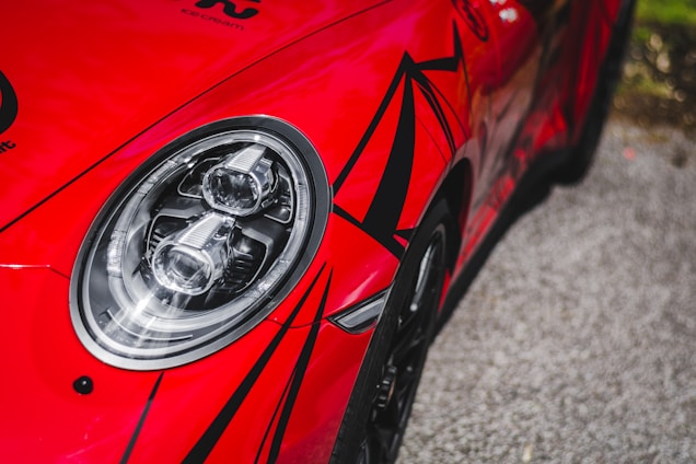 A close-up of a vibrant red sports car featuring a sleek headlight with intricate light fixtures. The car's body is adorned with bold black geometric patterns, accentuating its dynamic design. The image also shows a portion of a gray asphalt road in the background.