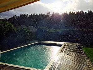 A swimming pool is surrounded by a wooden deck, with droplets of water visible in the air. The sun is shining brightly through a partly cloudy sky, casting reflections on the water. In the background, a dense line of trees provides a natural border.